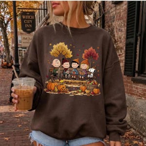May include: A dark brown crewneck sweatshirt featuring a graphic print of the Peanuts characters sitting on a bench surrounded by autumn trees, pumpkins, and falling leaves. The design includes Snoopy, Charlie Brown, and friends in fall attire.