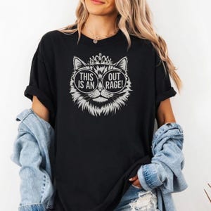 May include: Black t-shirt featuring a cat graphic with sunglasses and a crown. The graphic includes the text "THIS IS AN OUT RAGE!". The shirt has short sleeves and a relaxed fit, suitable for casual wear.