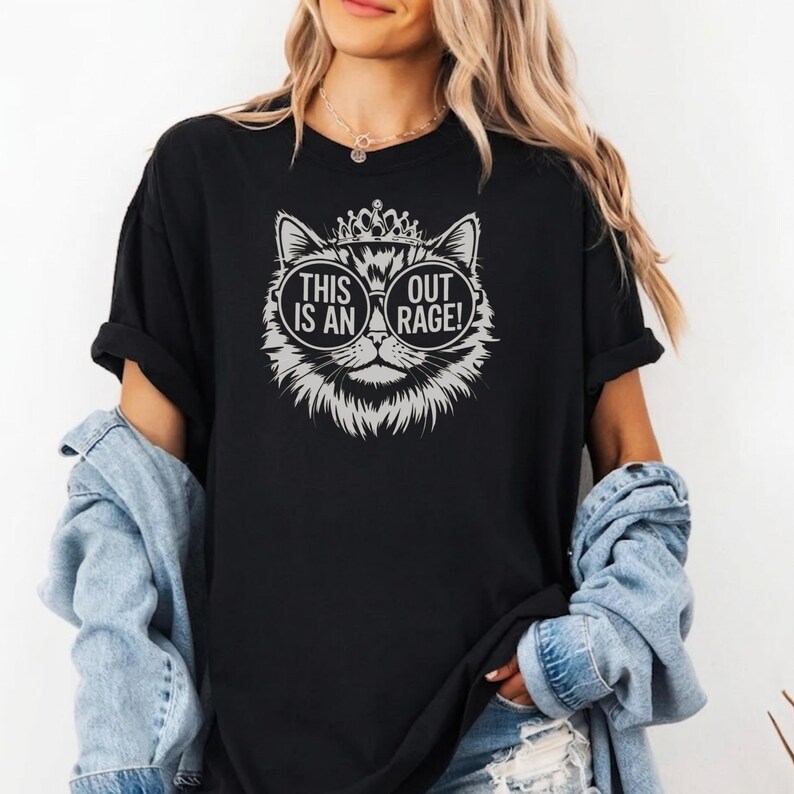 May include: Black t-shirt featuring a cat graphic with sunglasses and a crown. The graphic includes the text "THIS IS AN OUT RAGE!". The shirt has short sleeves and a relaxed fit, suitable for casual wear.