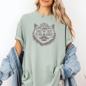 May include: A light green t-shirt featuring a cat graphic with sunglasses and a crown. The graphic includes the text "THIS IS AN OUT RAGE!". The shirt has short sleeves and a relaxed fit.