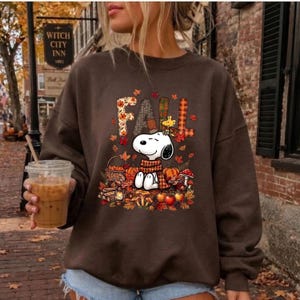 May include: A dark brown crewneck sweatshirt featuring a graphic print of the cartoon character Snoopy wearing a scarf. The text FALL is displayed above the character in a plaid pattern. The design includes autumn leaves, pumpkins, and apples.