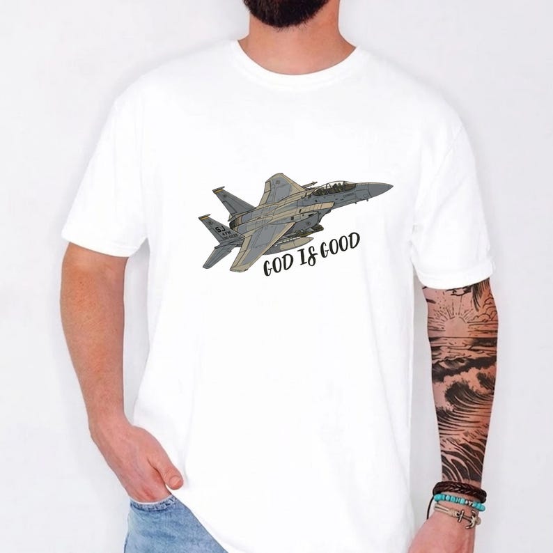 Pu&ograve; includere: T-shirt bianca con un'illustrazione dettagliata di un jet da combattimento grigio. Il testo "GOD IS GOOD" &egrave; stampato in nero. La maglietta &egrave; un classico girocollo.