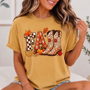 May include: A mustard yellow short-sleeve t-shirt featuring a graphic print of the word FALL. The letters are decorated with a black and white checkerboard pattern, a cactus motif, and a pair of western boots with floral and leopard print details.