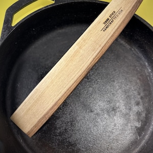 Peut inclure: Une poêle en fonte noire avec une spatule en bois clair. La spatule est rectangulaire et porte l'inscription "COOK STICK HANDCRAFTED USA". Le fond est jaune uni.