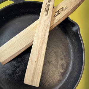 Peut inclure: Deux ustensiles de cuisine en bois clair sont posés sur une poêle en fonte noire. Le texte "COOK STICK HANDCRAFTED USA" est imprimé sur chaque ustensile. Le fond est jaune uni.