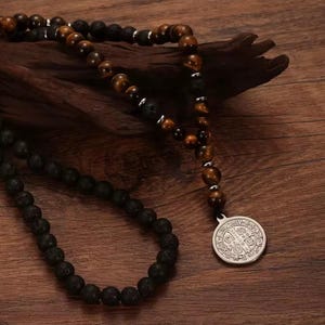 Könnte beinhalten: Eine Halskette mit schwarzen Lavasteinperlen, Tigeraugenperlen und einem silbernen Anhänger. Der Anhänger hat ein kompliziertes Design. Die Halskette wird auf einer Holzoberfläche präsentiert.