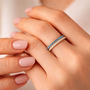 Op de afbeelding: Twee gouden ringen, één met blauwe edelstenen en de andere met witte edelstenen, worden aan een vinger gedragen. De ringen staan tegen een achtergrond van een hand met roze nagellak.