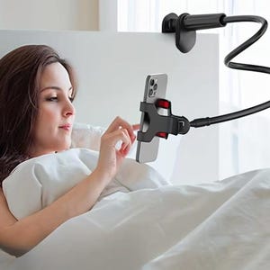 Könnte beinhalten: Ein schwarzer, flexibler Telefonhalter, der an einem Kopfteil befestigt ist. Der Halter hat eine Klemme zur Befestigung am Bettrahmen und eine Halterung zur Aufnahme eines Smartphones. Das Telefon ist silber und schwarz. Die Person im Bild benutzt das Telefon.