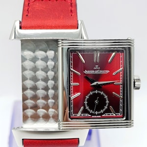 Könnte beinhalten: Eine rechteckige Jaeger-LeCoultre-Uhr mit rotem Zifferblatt und kleiner Sekunde. Die Uhr hat ein silbernes Gehäuse und ein rotes Lederarmband. Die Wörter "JAEGER-LECOULTRE" und "SWISS MADE" sind auf dem Zifferblatt zu sehen.