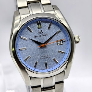 May include: A silver-toned Grand Seiko wristwatch with a light blue textured dial and a date window at the 3 o'clock position. The watch features black hands with an orange second hand and silver hour markers. The watchband is made of silver metal.
