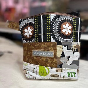 Może przedstawiać: Pikowana torba z tkaniny z zapięciem na zamek błyskawiczny. Torba ma wzór patchworkowy z motywami kwiatowymi, geometrycznymi i zwierzęcymi. Kolory obejmują brązowy, czarny, biały i zielony. Widoczna jest etykieta "Junco Studio".