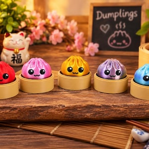 Könnte beinhalten: Fünf kleine, runde, bunte Knödel-Figuren mit lächelnden Gesichtern und großen schwarzen Augen, die jeweils auf einem beigen, kreisförmigen Sockel ruhen. Die Knödel sind rot, rosa, gold, lila und blau und in einer Reihe auf einer Holzoberfläche angeordnet.