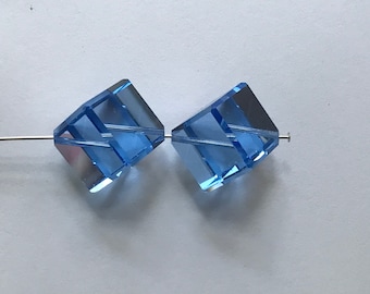 May include: Two light blue, faceted cube beads strung on a silver-colored wire. The beads are transparent, allowing light to pass through, and have a geometric design. The beads are likely for jewelry making.