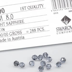 May include: A close-up of a package of 288 light sapphire Swarovski crystal beads. The beads are 8.0 mm in size and are made in Austria. The package is labeled "1st Quality" and "Satin".