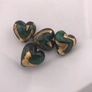 May include: Four heart-shaped beads with a dark green base and gold leaf accents. The beads are smooth and glossy, with a slight variation in the gold leaf pattern on each heart. They are arranged on a white surface.