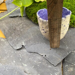 May include: A small, handmade ceramic cup with a speckled white exterior and a purple interior. The cup is being measured with a wooden ruler. The cup is approximately 5 cm tall. The background includes green moss and gray slate tiles.