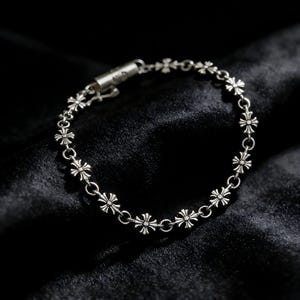 Può includere: Un braccialetto d'argento con una serie di piccoli ciondoli simmetrici a forma di croce. Il braccialetto è realizzato in un materiale metallico lucido ed è esposto su uno sfondo scuro e strutturato. La chiusura è visibile e il braccialetto sembra nuovo.