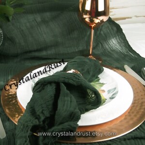 May include: A dark green linen napkin is folded and placed on a copper charger plate with a white dinner plate on top. A copper wine glass is in the background. The text "CrystalandRust" is visible on the napkin.