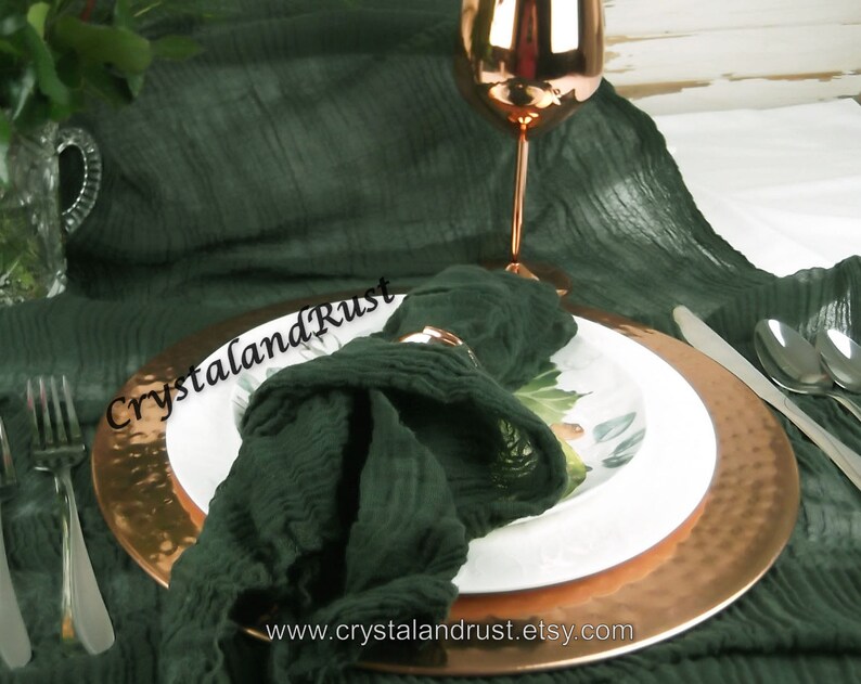 May include: A dark green linen napkin is folded and placed on a copper charger plate with a white dinner plate on top. A copper wine glass is in the background. The text "CrystalandRust" is visible on the napkin.