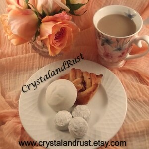 May include: A white plate with a pastry, four white chocolate covered candies, and a cup of coffee on a peach colored fabric background. The pastry is a swirl of dough with a brown filling. The candies are round and have a slightly rough texture. The coffee cup is white with a floral pattern and has a brown liquid inside. The fabric is soft and has a delicate texture.