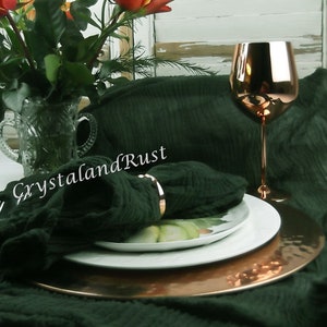 May include: A dark green linen tablecloth with a copper charger plate and a white plate on top. A copper wine glass sits on the table next to the plates. A bouquet of orange and red lilies sits in a clear glass vase in the background. The text "CrystalandRust" is visible on the tablecloth. The text "www.crystalandrust.etsy.com" is visible at the bottom of the image.