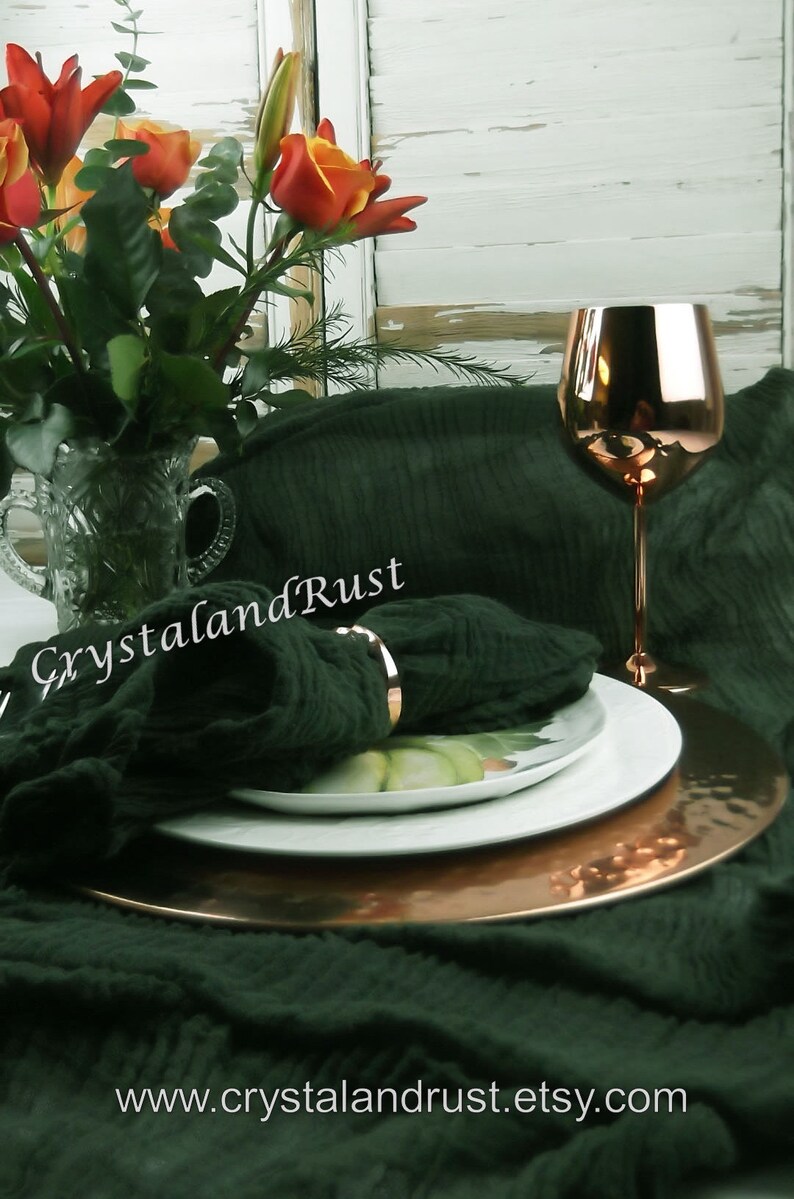 May include: A dark green linen tablecloth with a copper charger plate and a white plate on top. A copper wine glass sits on the table next to the plates. A bouquet of orange and red lilies sits in a clear glass vase in the background. The text "CrystalandRust" is visible on the tablecloth. The text "www.crystalandrust.etsy.com" is visible at the bottom of the image.