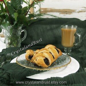 May include: A close-up of a plate of pastries with a glass of coffee on a green tablecloth. The pastries are filled with a dark filling and have a golden brown crust. The coffee is in a clear glass with a handle and is filled to the top.