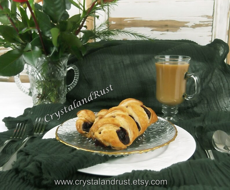 May include: A close-up of a plate of pastries with a glass of coffee on a green tablecloth. The pastries are filled with a dark filling and have a golden brown crust. The coffee is in a clear glass with a handle and is filled to the top.