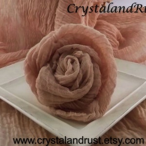 May include: A rose-colored fabric rosette on a white square plate. The fabric is soft and crinkled, creating a delicate and romantic look. The fabric is arranged in a circular shape, resembling a flower.