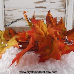 May include: A bunch of artificial fall leaves in shades of orange, yellow, and red, arranged on a white fluffy surface. The leaves are made of fabric and have a realistic appearance.