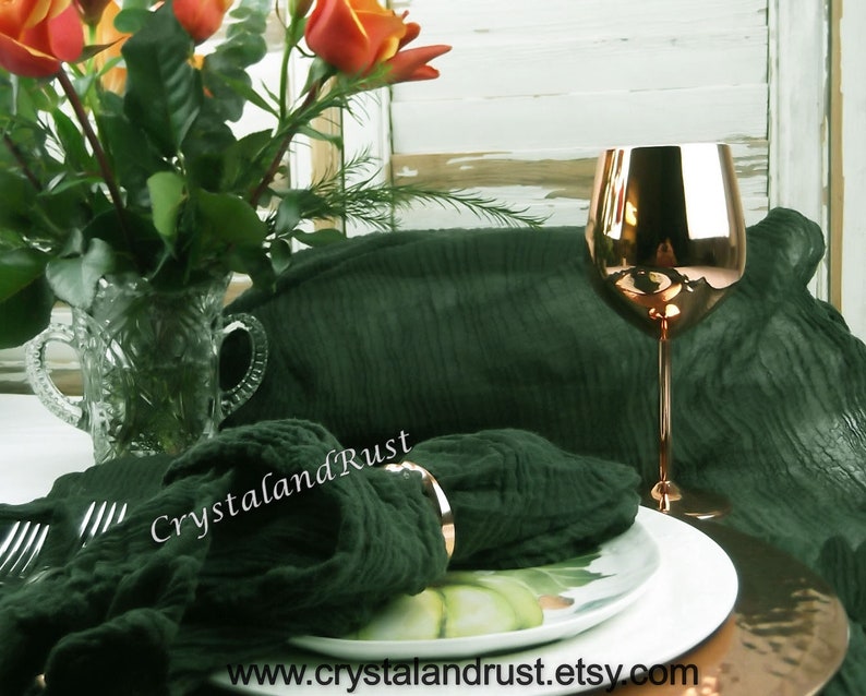 May include: A dark green linen napkin with a silver napkin ring on a white plate with a green and white design. A copper wine glass sits on the plate. The text "CrystalandRust" is visible on the napkin.