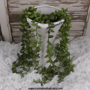 May include: A white ceramic vase with a green string of pearls plant hanging over the edge. The vase is sitting on a white ruffled fabric surface. The text "CrystalandRust" is visible on the vase.