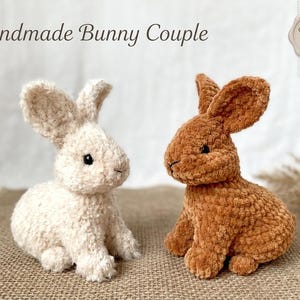 Puede incluir: Dos peluches de conejito hechos a mano. Uno es blanquecino, el otro es marrón. Los conejitos están sentados sobre una superficie de arpillera. El texto "Handmade Bunny Couple" está encima de los conejitos. Una pequeña etiqueta dice "Crafted with Love".