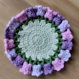 以下が含まれることがあります： 手作りの、花柄デザインの丸いかぎ針編みのコースター。コースターは、緑の葉と紫とピンクの花の形をした装飾で囲まれた、スパイラル模様の淡い緑色の中心部を持っています。コースターの直径は約13cmです。