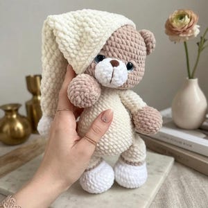 Puede incluir: Un oso de peluche de ganchillo hecho a mano con un gorro de dormir color crema y un pijama a juego. El oso tiene un cuerpo marrón claro, extremidades color crema y ojos azules. El oso mide aproximadamente 25 cm de alto y es un juguete suave y tierno.