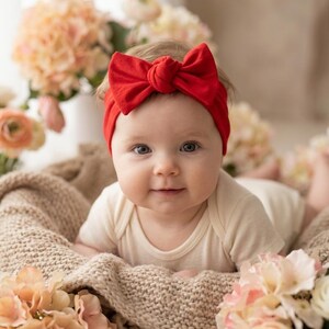 Könnte beinhalten: Ein Baby mit einem leuchtend roten Schleifen-Stirnband und einem cremefarbenen Body. Das Baby liegt auf einer beigen Strickdecke, umgeben von pfirsichfarbenen und cremefarbenen Blumen. Das Baby schaut direkt in die Kamera.