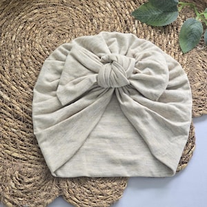 Könnte beinhalten: Eine hellbeige Turban-Mütze mit einem markanten, geknoteten Schleifendetail in der vorderen Mitte. Der Stoff hat eine weiche, melierte Textur mit sichtbaren Falten und Raffungen, die vom zentralen Knoten ausgehen und eine runde Form bilden.