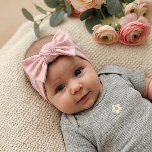 Könnte beinhalten: Ein Baby trägt ein hellrosa Knoten-Stirnband mit weißem Punktemuster und einen grauen, gerippten Kurzarm-Body mit einer kleinen, aufgestickten weißen Blume auf der Brust. Das Baby liegt in der Nähe eines Straußes aus hellrosa Blumen.