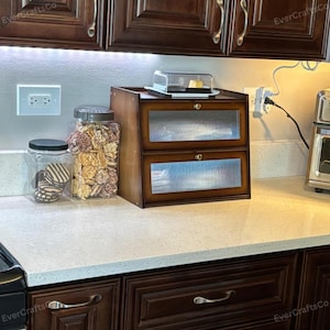 May include: A wooden bread box with two glass-fronted drawers, a clear plastic container, and two glass jars filled with cookies and snacks. The bread box is on a white countertop in a kitchen.