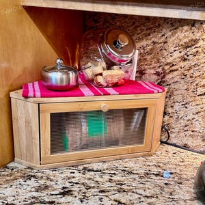 May include: A bamboo bread box with a glass door and a wooden knob. A red and white striped cloth sits on top, with a small silver bowl and a glass jar filled with baked goods. The bread box is on a countertop.
