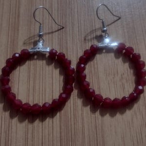 May include: A pair of hoop earrings featuring a deep red, faceted bead design. The beads are strung to create a circular shape, with silver-coloured hooks. The earrings are displayed on a wooden surface.