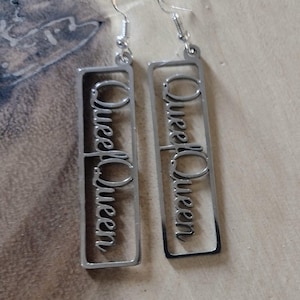 May include: A pair of silver-toned dangle earrings featuring a rectangular frame with the word Queen written in a cursive script inside each frame. The earrings are attached to standard silver-toned metal earring hooks for pierced ears.
