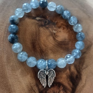 Könnte beinhalten: Ein Armband mit hellblauen, runden Edelsteinen. Es hat einen silberfarbenen Anhänger in Form von Engelsflügeln. Das Armband liegt auf einer Holzoberfläche.