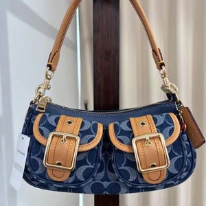 May include: A blue denim shoulder bag with two front pockets, tan leather accents, and gold-tone hardware. The bag features a top zip closure, a leather handle, and a small tag. The Coach logo is visible on a tag.