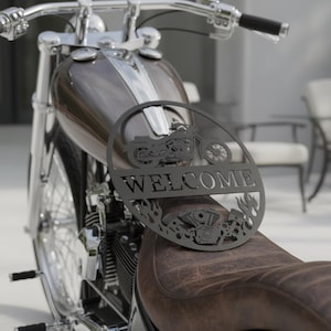 Könnte beinhalten: Schwarzes Metall-Willkommensschild mit Motorrad-Design, Flammen und Motordetails. Das Schild liegt auf dem Sitz eines braunen Motorrads. Das Motorrad hat einen Chromlenker und einen braunen Tank.