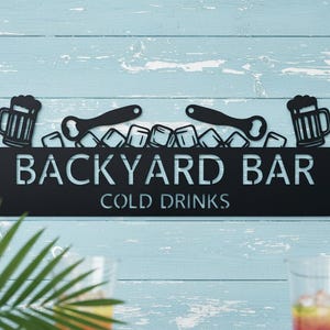 Könnte beinhalten: Schwarzes Metallschild mit der Aufschrift "BACKYARD BAR COLD DRINKS". Es zeigt Bierkrüge, Flaschenöffner und Eiswürfel. Das Schild ist auf einem hellblauen Holzgrund montiert, ideal für eine Gartenbar.