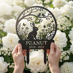 Könnte beinhalten: Schwarzer Metall-Gartenstecker mit kreisförmigem Design mit einem Kaninchen, Vögeln, Schmetterlingen und floralen Elementen. Der Stecker trägt die Aufschrift "PEANUT 2018-2024". Der Hintergrund ist mit weißen Blumen gefüllt.