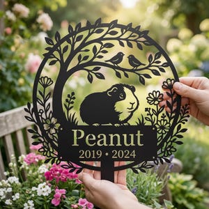 Könnte beinhalten: Ein schwarzer, kreisförmiger Gartenstecker aus Metall mit der Silhouette eines Meerschweinchens, zwei Vögeln auf einem Ast und floralen Verzierungen. Der Text "Peanut" steht mittig über den Jahren "2019 • 2024" auf einer rechteckigen Plakette.