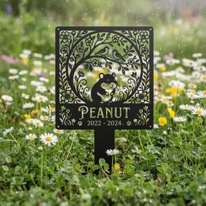 Könnte beinhalten: Schwarzer Metall-Gartenstecker mit dekorativem Ausschnitt-Design. Das Design zeigt einen Hamster, Vögel, Blumen und den Namen "PEANUT" mit den Jahren "2022-2024". Der Stecker steht in einem Garten mit Gänseblümchen.