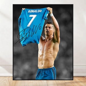 May include: A framed print featuring a soccer player holding up a blue jersey. The jersey displays the name "RONALDO" and the number "7". The player is shirtless, wearing blue shorts, and the background is a blurred black and white image.